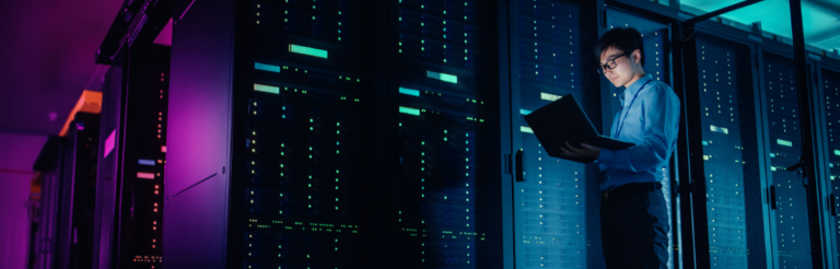 In Data Center - Male IT Technician Running Maintenance Programme on a Laptop, Controls Operational Server Rack Optimal Functioning. Modern High-Tech Operational Super Computer in Neon Colours, Lights
