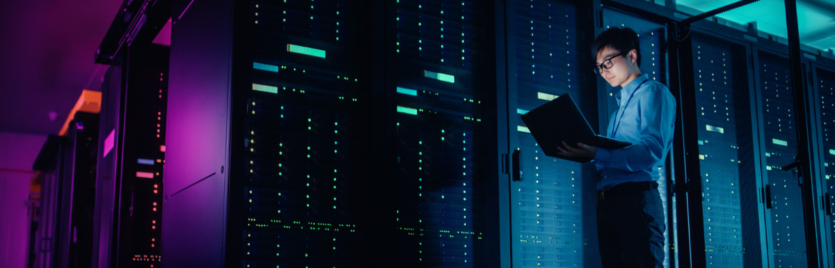 In Data Center - Male IT Technician Running Maintenance Programme on a Laptop, Controls Operational Server Rack Optimal Functioning. Modern High-Tech Operational Super Computer in Neon Colours, Lights